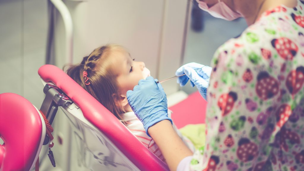 Bir pedodontist, diş kliniğinde küçük bir çocuğun dişlerini nazikçe muayene ediyor ve çocuk diş sağlığı kontrolü yapıyor.